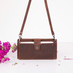 Puede incluir: Un bolso bandolera rectangular de cuero marrón que presenta una solapa frontal con cierre de broche de metal, una cremallera superior y una correa de hombro delgada y ajustable sujeta con herrajes de metal plateado.