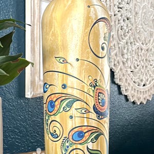 May include: A decorative, hand-painted bottle with a gold base and peacock feather design. The bottle has a white rope detail at the neck and is displayed on a wooden surface. The bottle is a unique piece of home decor.