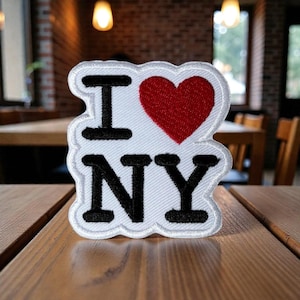 Puede incluir: Parche bordado blanco con el texto "I NY" en negro y un corazón rojo. El parche está sobre una superficie de madera, con un fondo borroso de restaurante.