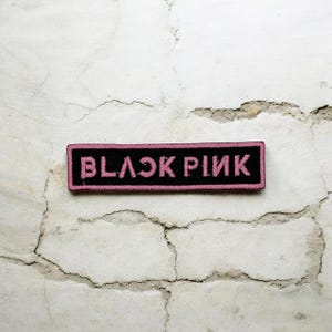 Peut inclure: Un écusson rectangulaire noir avec les mots "BLACK PINK" brodés en rose. L'écusson a une bordure rose et est posé sur un fond blanc texturé.