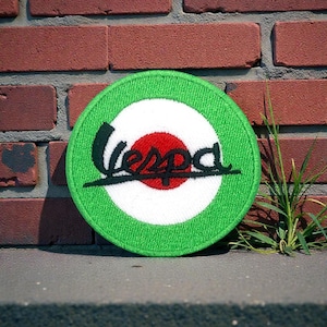 May include: A round, green and white patch with the word "Vespa" in black script. The patch has a red center and is set against a brick wall and concrete surface.