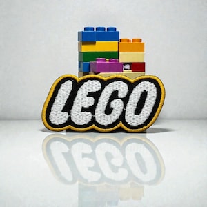 May include: A black and white LEGO patch with a yellow border sits in front of a stack of colourful LEGO bricks. The bricks are blue, yellow, orange, green, purple, and red. The patch and bricks are reflected on a white surface.