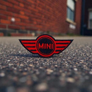Puede incluir: Un parche bordado en rojo y negro con la palabra "MINI" en un óvalo blanco, con alas rojas. El parche está colocado sobre una superficie gris texturizada, con una pared de ladrillos borrosa en el fondo.