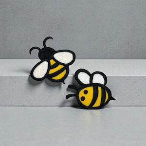 May include: Two embroidered bee patches. One bee is angled upward, the other is angled downward. Both bees have black bodies, yellow and black striped abdomens, and white wings. The patches are on a gray surface.