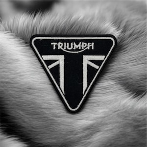Può includere: Toppa Triumph in bianco e nero a forma triangolare. La toppa presenta il logo Triumph e un design stilizzato della bandiera Union Jack. La toppa è posta su uno sfondo di morbida e strutturata pelliccia grigia.