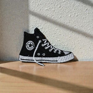 Puede incluir: Un parche bordado en blanco y negro con forma de zapatilla alta. El parche presenta un logotipo de estrella blanca y cordones blancos. La zapatilla se encuentra sobre una pared blanca texturizada y una superficie de madera.
