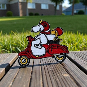 以下が含まれることがあります： 赤いスクーターに乗ったスヌーピーとウッドストックの刺繍パッチ。スヌーピーは赤いヘルメットとスカーフを着用し、ウッドストックは彼の後ろに座っています。スクーターは黒と黄色の車輪があり、木製のデッキと緑の草を背景にしています。