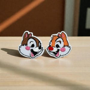 以下が含まれることがあります： 漫画のキャラクターの刺繍パッチが2つ。片方は茶色の毛皮、もう片方はオレンジ色。どちらも白い顔、ピンクの頬で、舌を出して笑っています。パッチは黒で縁取られています。衣類やアクセサリーに最適です。