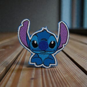 Puede incluir: Un parche bordado azul con el personaje Stitch de la película Lilo & Stitch. El parche tiene un borde blanco, ojos negros grandes, orejas moradas y una nariz azul. El parche está sobre una superficie de madera.