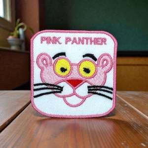 Op de afbeelding: Geborduurde patch met het stripfiguur Pink Panther. De vierkante patch heeft een witte achtergrond en een roze rand. Het gezicht van de Roze Panter staat in het midden, met gele ogen, een rode neus en zwarte snorharen. De tekst "PINK PANTHER" staat bovenaan.