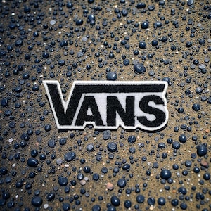 Può includere: Una toppa Vans in bianco e nero con il nome del marchio in grassetto. La toppa ha un bordo bianco ed è posizionata su una superficie strutturata ricoperta di piccoli ciottoli blu scuro.