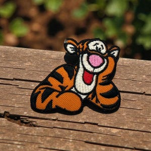 May include: Embroidered patch featuring Tigger from Winnie the Pooh. The patch has orange and black stripes, a pink nose, and a smiling expression. The patch is on a wooden surface.