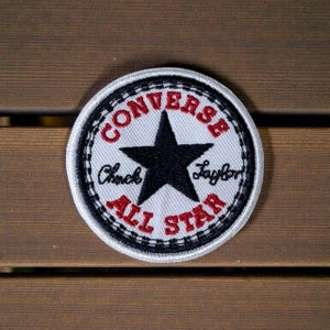 Puede incluir: Parche circular con el logotipo de Converse All Star. El parche tiene un fondo blanco con una estrella negra en el centro. Las palabras "CONVERSE" y "ALL STAR" son rojas, con "Chuck Taylor" en negro.