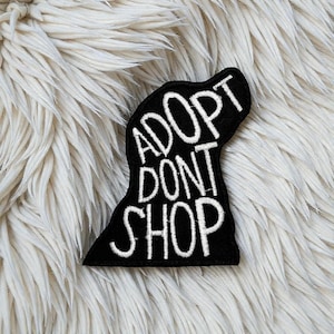 Op de afbeelding: Een zwarte patch in de vorm van een hondenhoofd met de woorden "ADOPT DONT SHOP" in het wit geborduurd. De patch is geplaatst tegen een pluizige, witte achtergrond.