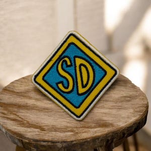 Op de afbeelding: Een vierkantige patch met een turquoise blauwe ruit met de gele letters "SD". De ruit is geel en wit omlijnd. De patch ligt op een houten oppervlak.