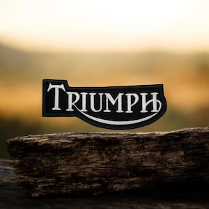 Op de afbeelding: Een zwart-wit geborduurde patch met het woord "TRIUMPH" in een gestileerd lettertype. De patch heeft een rechthoekige vorm met een gebogen onderrand en een unieke bovenrand. De achtergrond is wazig, met een houten oppervlak op de voorgrond.
