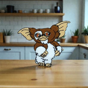 May include: Embroidered patch of a Gremlin character in brown and white, with large ears and orange eyes. The patch has a detailed, stitched design, suitable for clothing or accessories. The patch is displayed on a wooden surface.