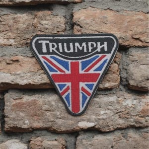 Könnte beinhalten: Ein dreieckiger Triumph-Aufnäher mit weißem Schriftzug auf schwarzem Grund. Der Aufnäher zeigt ein Union Jack-Design in Rot, Weiß und Blau. Der Aufnäher befindet sich vor einer Backsteinmauer.