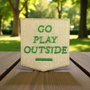 Puede incluir: Un parche bordado beige con las palabras "GO PLAY OUTSIDE" en verde. El parche tiene forma de escudo y está sobre una superficie de madera. El fondo es una vista borrosa de un parque.