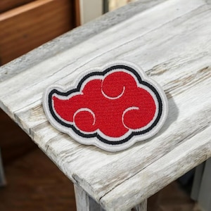Op de afbeelding: Een rode wolk-vormige patch met een zwarte omtrek en witte rand. De patch is geborduurd met een gedetailleerd ontwerp en is waarschijnlijk voor kleding of accessoires. De patch is geplaatst op een verweerd houten oppervlak.