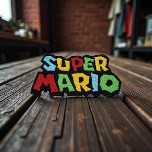 Könnte beinhalten: Gestickter Aufnäher mit dem Schriftzug "SUPER MARIO" in bunten Farben. Der Aufnäher hat einen schwarzen Rand und liegt auf einer rustikalen Holzoberfläche. Die Buchstaben sind rot, gelb, grün und blau.