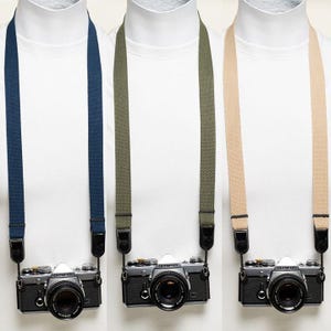 May include: Three Olympus cameras, each featuring a different coloured woven fabric neck strap. The straps are navy blue, olive green, and beige. Each camera is silver and black with a lens attached, suspended by black connectors on the straps.