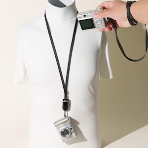 May include: A black camera neck strap featuring a quick-release buckle mechanism. The strap is shown attached to a silver compact digital camera hanging against a white mannequin torso, with an additional silver camera held nearby.