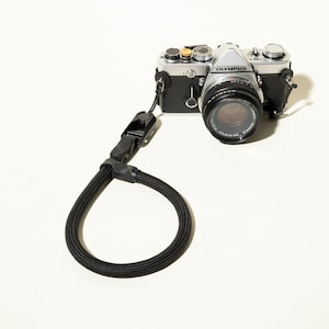 May include: A silver and black Olympus OM-1 film camera with a lens attached, featuring a thick, black, braided rope wrist strap connected to the side lug. The camera body has various dials, a shutter button, and the Olympus brand name printed on top.
