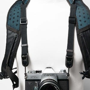 May include: A silver and black Canon FTb film camera with a 50mm lens is attached to a black camera harness. The harness features teal mesh padding on the shoulder straps and adjustable black nylon webbing with metal clips.