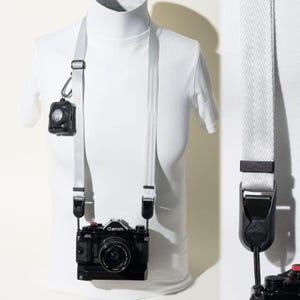 May include: A light grey camera strap with black hardware attachments, connected to a black Canon A-1 film camera. The strap features a sliding adjustment buckle and a small black carabiner holding a circular black accessory on the side strap.
