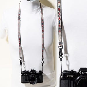 May include: A black Canon AE-1 camera attached to a woven camera strap featuring a beige background with a repeating geometric pattern in red, blue, and black. The strap includes black plastic hardware, adjustable sliders, and swivel clips.