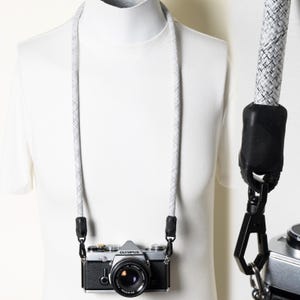 May include: A camera strap made of a white and gray woven rope material. The strap features black rubber connectors at the ends, which attach to a silver and black Olympus camera via black metal swivel clips. The rope has a textured pattern.