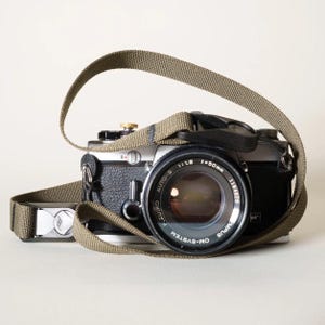 May include: A silver and black Olympus OM-System camera with a 50mm f/1.8 lens and a khaki-colored woven camera strap. The camera body features a textured black grip, a silver top plate, and a gold-colored shutter button on the top right side.