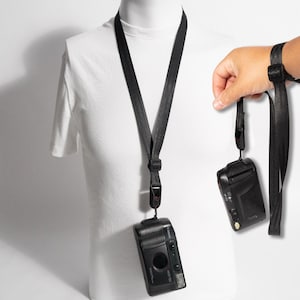 May include: A black camera strap made of smooth, durable webbing material. The strap features an adjustable sliding buckle and a secure attachment point for a camera. One end is shown connected to a black Canon camera, while the other end is held.