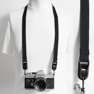 Könnte beinhalten: Ein schwarzer Kameragurt mit Schnellverschlüssen, befestigt an einer silber-schwarzen Canon FTb Filmkamera. Der Gurt besteht aus gewebtem Stoff und verfügt über schwarze Kunststoffbeschläge mit kleinen roten Akzenten an den Verbindungen.