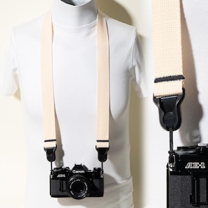 May include: A cream-coloured woven camera strap attached to a black Canon AE-1 film camera. The strap features black stitching details at the ends where it connects to the camera body via black plastic quick-release connectors and thin cords.