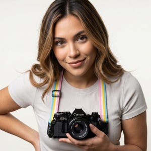 May include: A black Canon AE-1 35mm film camera with a lens attached, suspended by a colorful camera strap featuring vertical stripes in pink, yellow, white, and light blue, worn over a plain light gray short-sleeved crew neck shirt.