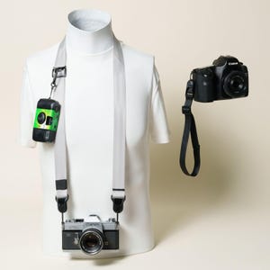 May include: A light gray camera strap worn on a white mannequin torso, featuring a silver Canon film camera attached at the bottom and a small green and black disposable camera clipped to the side. A black Canon DSLR camera hangs separately.