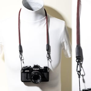 May include: A black Canon AE-1 camera with a 50mm lens attached to a dark red and black woven rope camera strap. The strap features black rubber connectors and metal swivel clips that attach to the camera body, providing a secure hold.