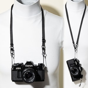 May include: A black Canon AE-1 35mm film camera with a 50mm lens attached, suspended by a thin black adjustable camera strap featuring metal swivel clips. The camera body displays the Canon logo and AE-1 model text on the front housing.