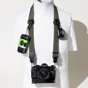 May include: An olive green camera strap attached to a black Canon AE-1 camera. Two small, portable cameras are clipped to the strap using black carabiners. One small camera is black and green, while the other is solid black. The strap is worn on a white mannequin.