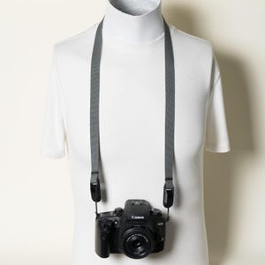 May include: A black Canon EOS digital camera with a lens attached, suspended from a plain gray fabric neck strap. The camera is positioned against a white t-shirt worn by a mannequin, featuring the Canon logo and EOS branding on the front body.