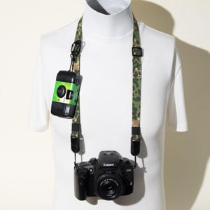 May include: A camouflage patterned camera strap featuring a black plastic carabiner attachment. A black Canon EOS DSLR camera is suspended from the strap, while a small green and black disposable camera hangs from the carabiner clip.