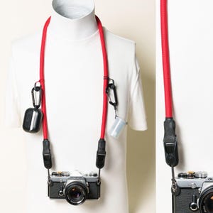 May include: A bright red camera neck strap featuring black hardware, two black carabiners, and black leather connectors. The strap is attached to a silver and black Olympus camera, with a black earbud case and a small translucent container clipped on.