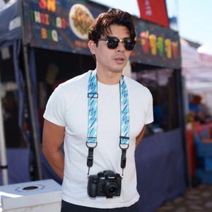 May include: A black digital camera hangs from a light blue camera strap featuring a repeating pattern of purple and white zig-zag lines. The strap is adjustable with black plastic hardware and connects to the camera body with black leather tabs.