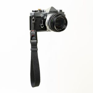 May include: A silver and black Olympus film camera with a black lens attached. A black fabric wrist strap is connected to the camera body via a quick-release mechanism featuring a small red accent. The camera body has a textured grip.