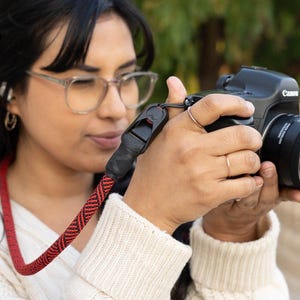 Può includere: Una fotocamera reflex digitale Canon nera con obiettivo, dotata di una tracolla in corda intrecciata rossa e nera. La tracolla presenta un connettore rapido in plastica nera e un motivo testurizzato lungo tutto il cordino.