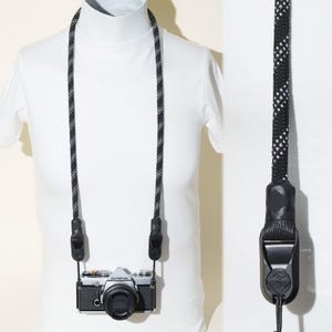 May include: A black and white patterned rope camera strap attached to a silver and black Olympus camera. The strap features a braided texture with reflective white accents and black plastic connectors at the attachment points.