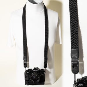 May include: A black camera strap with a woven textured pattern attached to a black Canon A-1 film camera. The strap features dark hardware connectors and a durable fabric construction, shown hanging on a white mannequin torso for display.