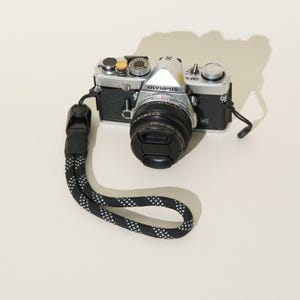 May include: A silver and black Olympus OM-1 camera with a lens cap attached. The camera features a textured black grip, silver top plate with dials, and a black and white patterned rope wrist strap connected to the side lug.